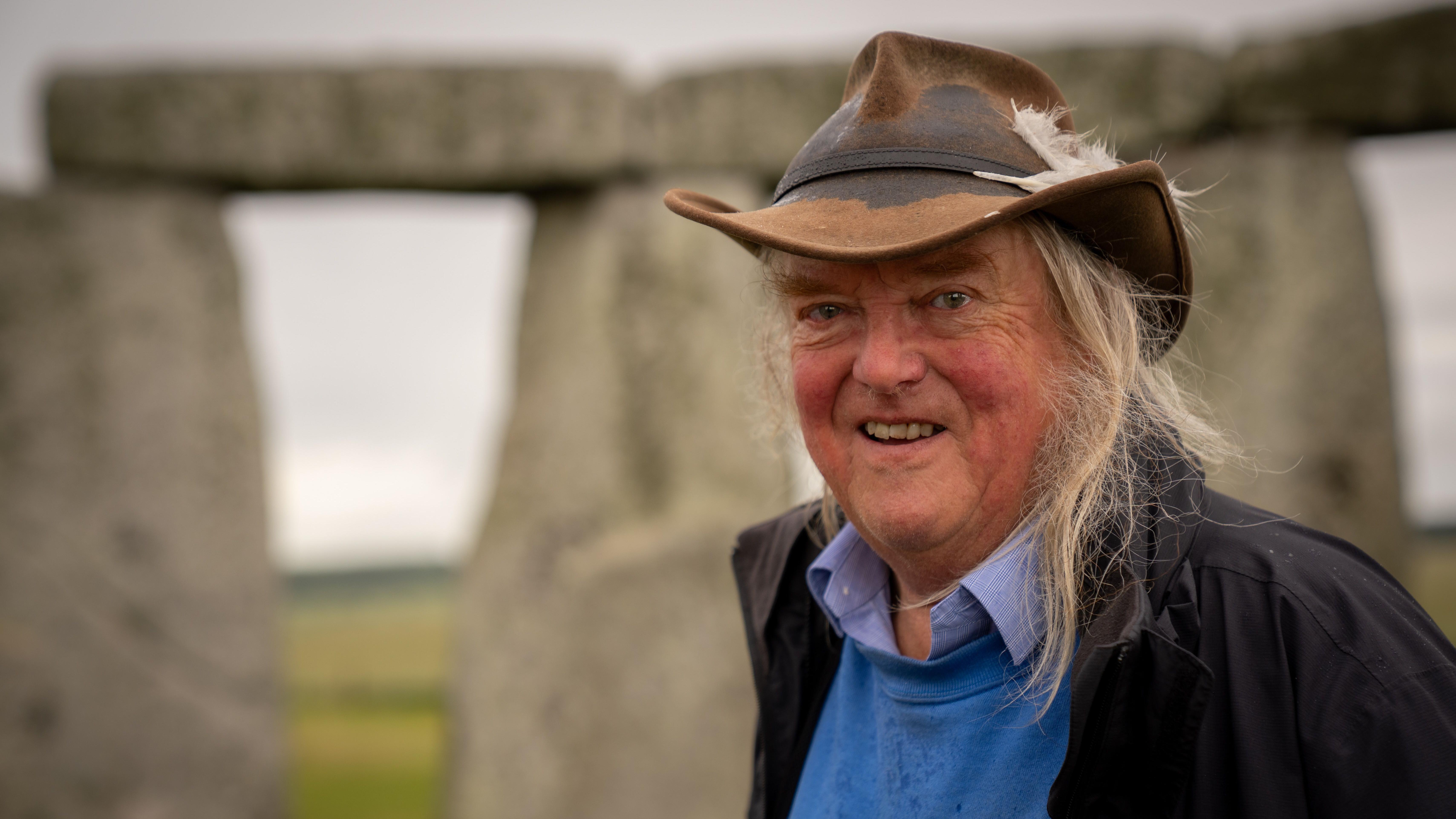 O arque&oacute;logo Phil Harding, que costumava aparecer no programa &ldquo;Time Team&rdquo; do Channel 4, usando seu chap&eacute;u de marca registrada com duas penas enfiadas na faixa do lado esquerdo. Seus longos cabelos louros est&atilde;o pendurados em cada lado de seu rosto muito caracter&iacute;stico. Ele est&aacute; vestindo uma camisa azul e um moletom, com um casaco aberto por cima. Atr&aacute;s dele, fora de foco, o c&iacute;rculo externo de Stonehenge &eacute; vis&iacute;vel e, al&eacute;m dele, a grama da plan&iacute;cie de Salisbury. 