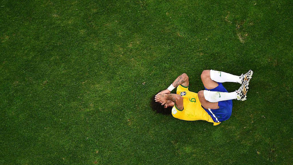 A imagem mostra um jogador da sele&ccedil;&atilde;o brasileira de futebol ca&iacute;do no gramado, encolhido e com as m&atilde;os cobrindo o rosto, sugerindo dor ou frustra&ccedil;&atilde;o ap&oacute;s um lance. Registrada de um &acirc;ngulo a&eacute;reo, a fotografia destaca o contraste entre a figura do atleta e a ampla &aacute;rea verde do campo, refor&ccedil;ando a sensa&ccedil;&atilde;o de isolamento no momento.