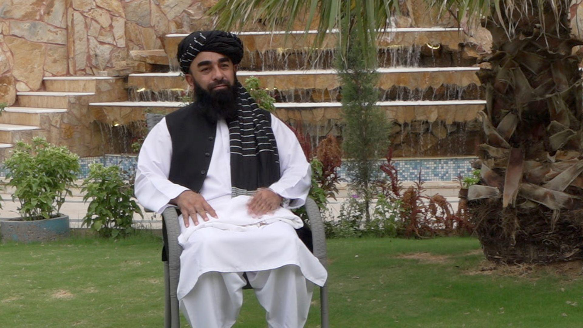A man in traditional white Afghan tunic and trousers with a black waistcoat and headdress sits in a chair on a lawn. Behind him are a large fountain, neatly laid stone steps, shrubs and a tree. 