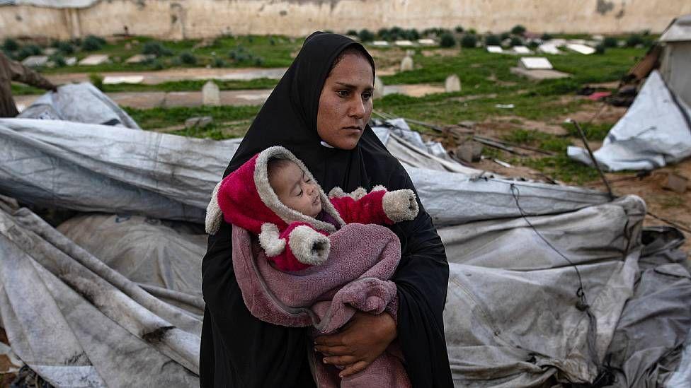 Una madre con un bebé en brazos junto a una tienda de campaña destruida.