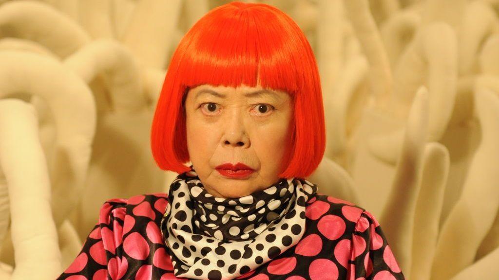 Yayoi Kusama con su caracteristico pelo rojo y su ropa de puntos de colores, en este caso una camisa negra con puntos rojos y una pañoleta blanco y negro con puntos de esos dos colores.