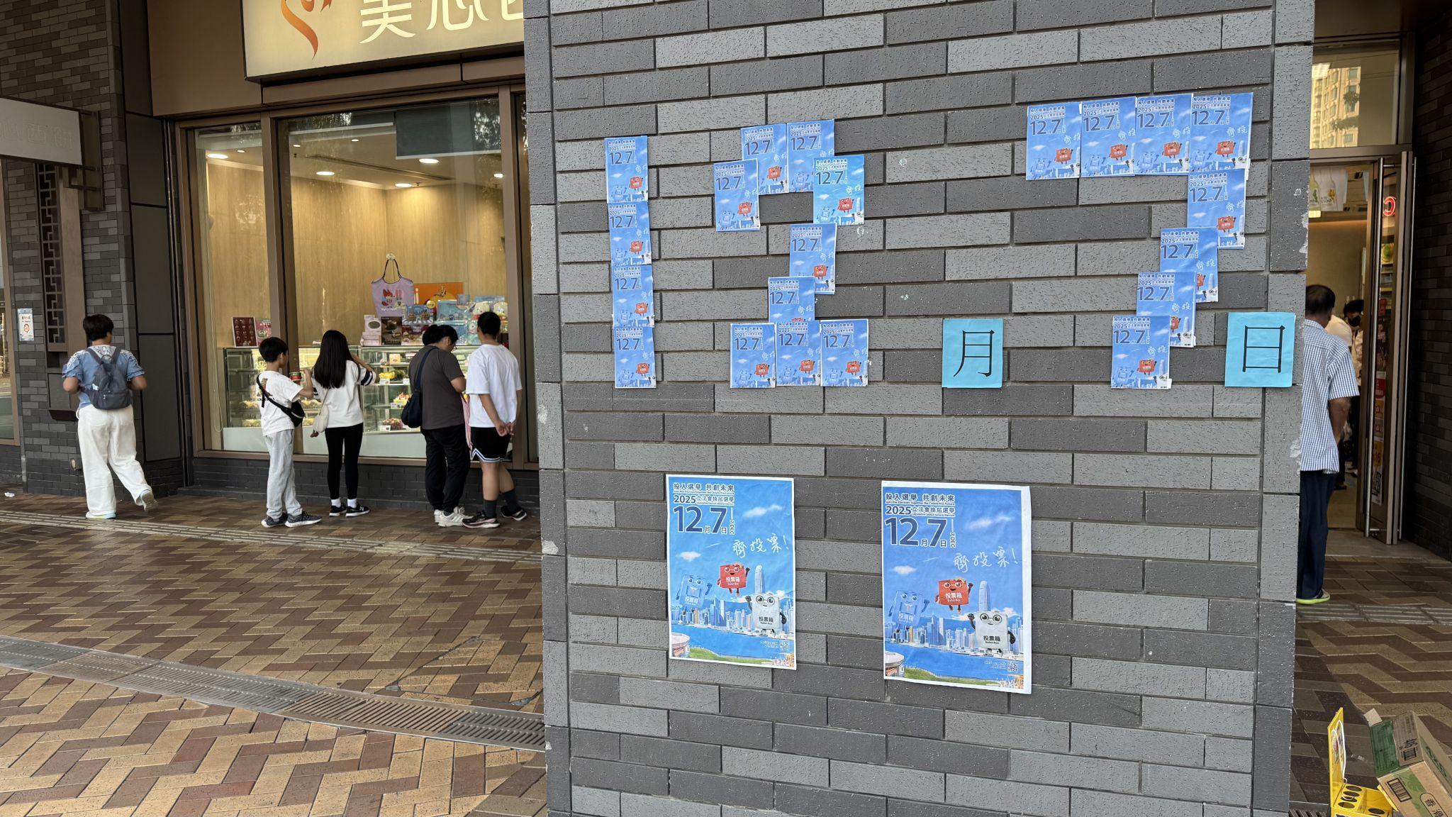 香港元朗洪水橋洪福邨商場一面牆上貼上了以小張12月7日立法會選舉投票官方宣傳海報堆砌成的「12月7日」字樣（BBC中文圖片16/11/2025）