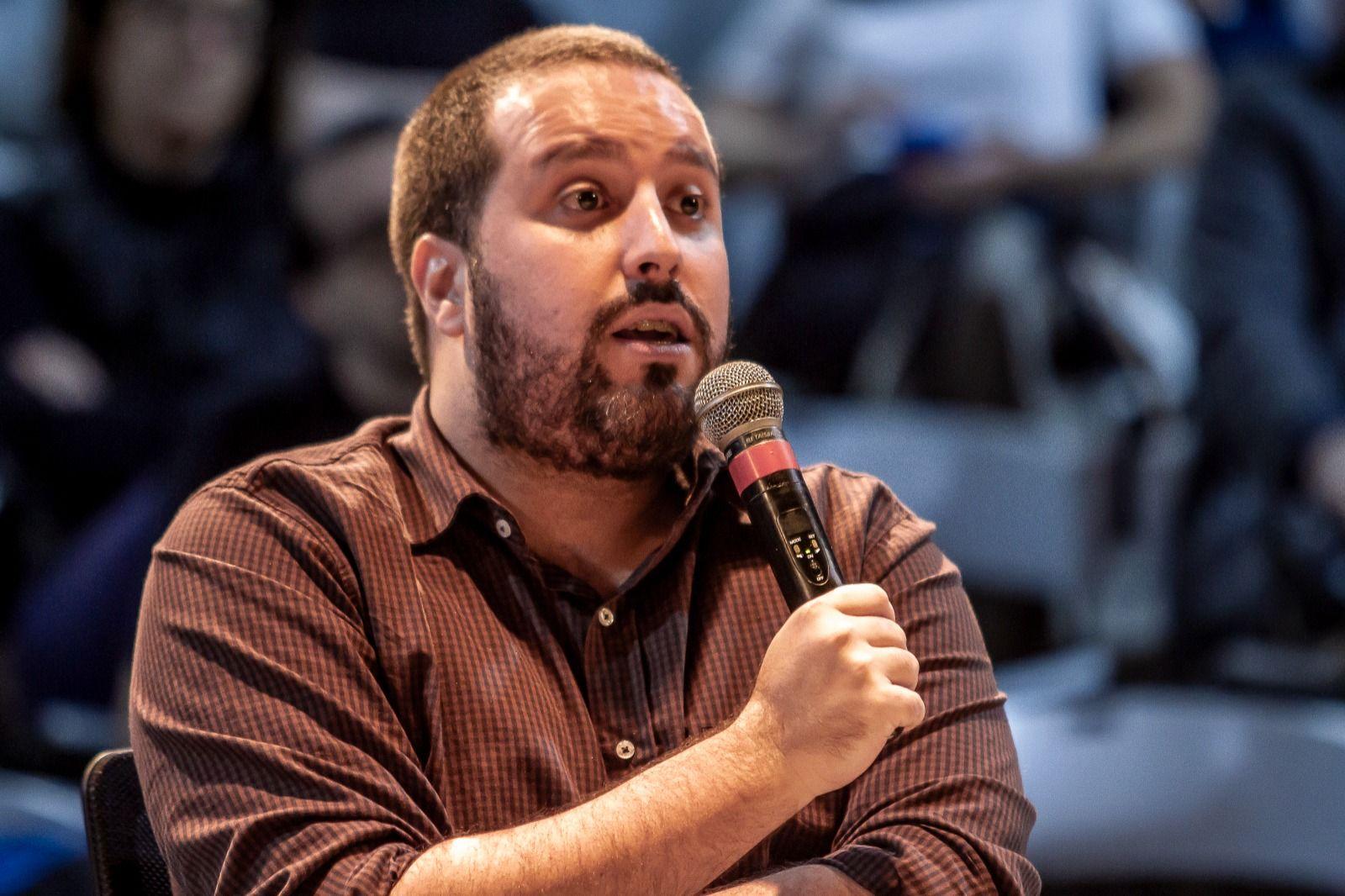 Rafael Soares sentado enquanto fala ao microfone, com feição atenta e séria