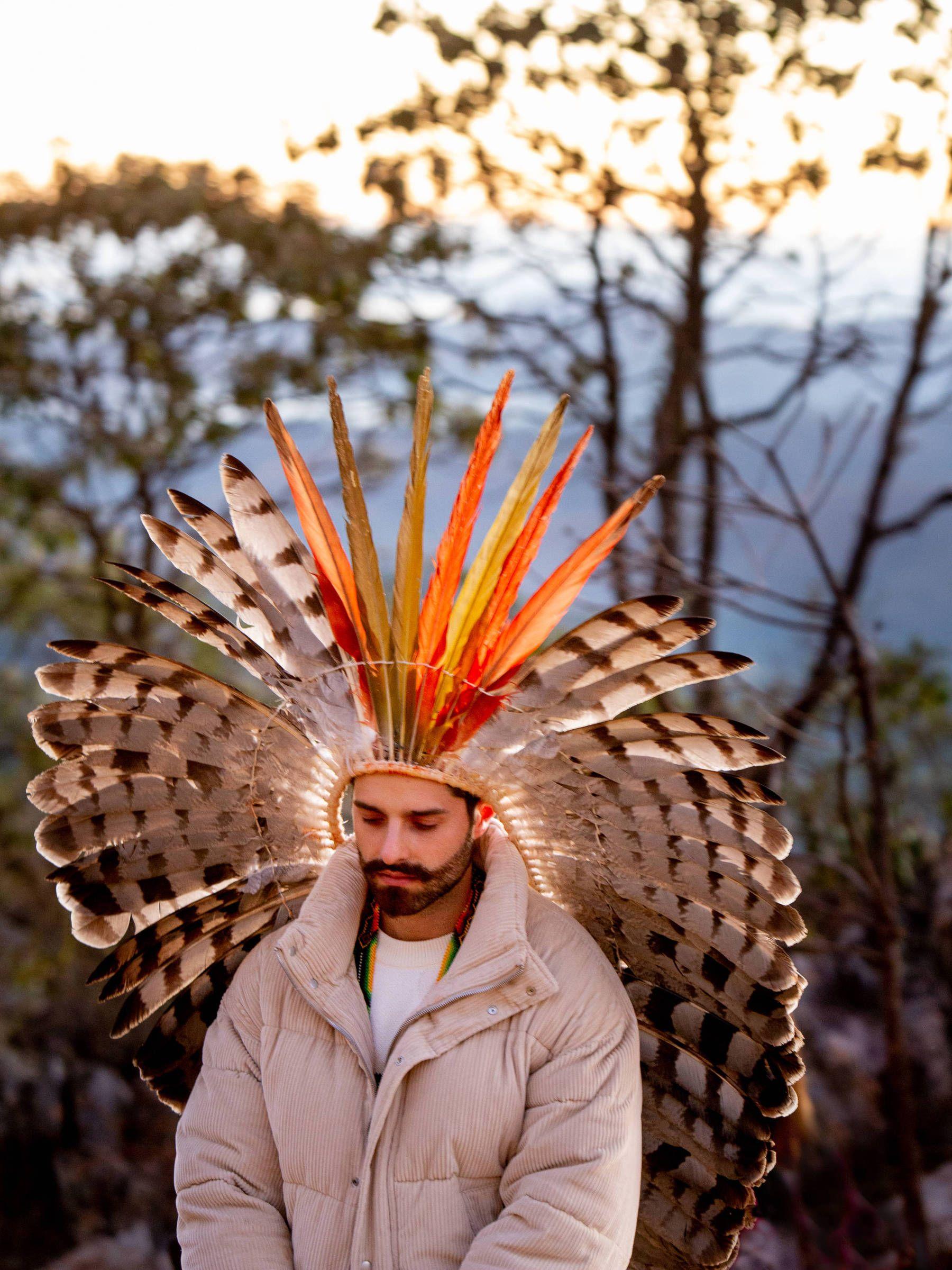 Alok de olhos fechados em meio &agrave; natureza, usando um cocar de grandes penas em tons de branco, preto, laranja e amarelo, que ocupa quase toda a composi&ccedil;&atilde;o. Vestido com um casaco acolchoado bege, ele aparece em uma postura contemplativa.
