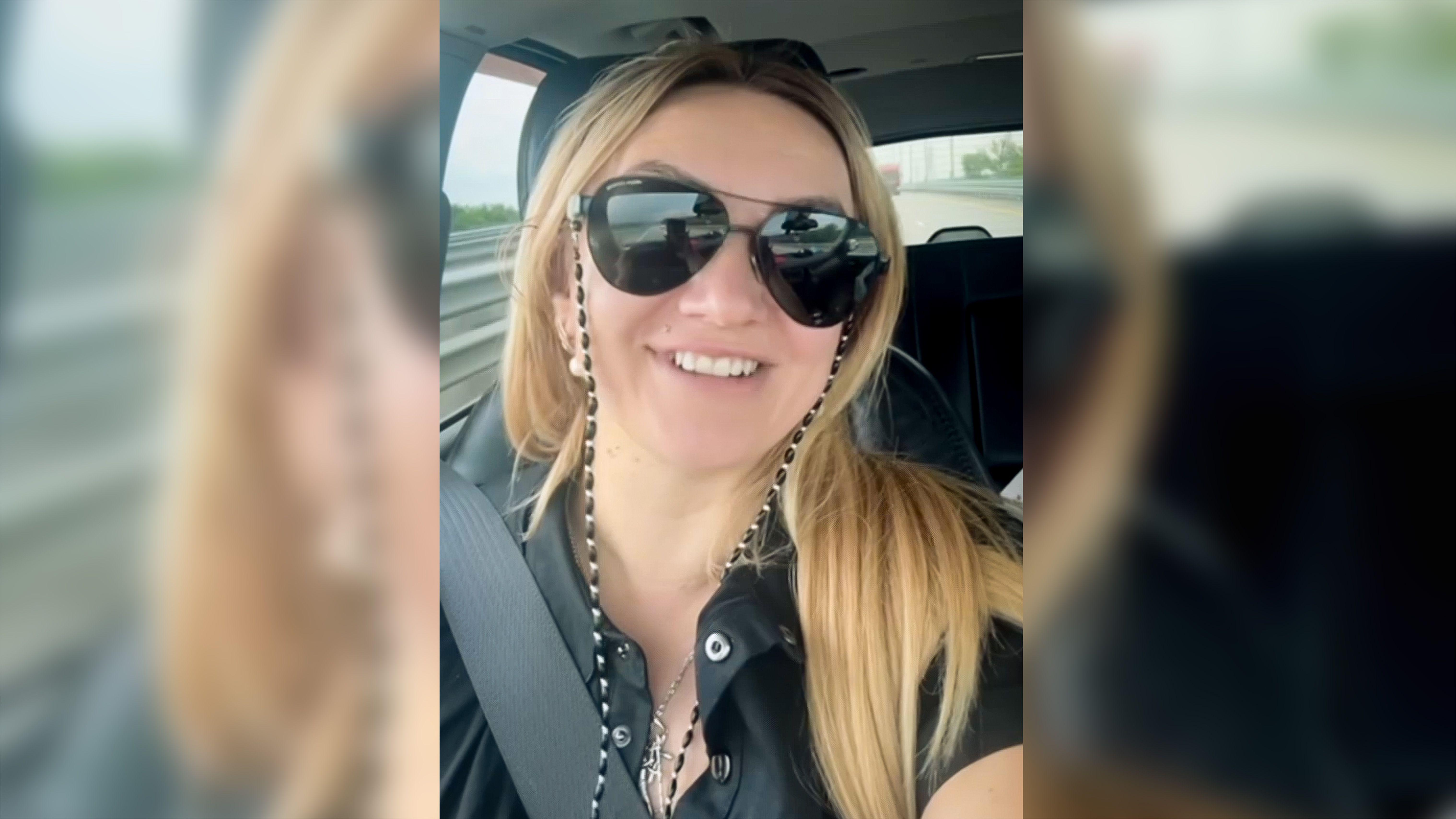 Polina Azarnik gleda pravo u kameru dok sedi u vozilu. Ima dugu plavu kosu, nosi velike naočare za sunce i crnu majicu i osmehuje se