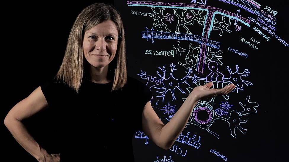 A professora Noelia Valle com camiseta preta e cabelos loiros, em frente a uma lousa com desenhos coloridos sobre biologia