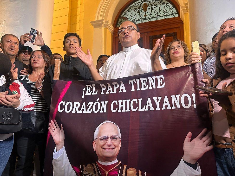 Un sacerdote rodeado de personas y frente a una pancarta con la foto del Papa León XIV que dice: El Papa tiene corazón chiclayano.