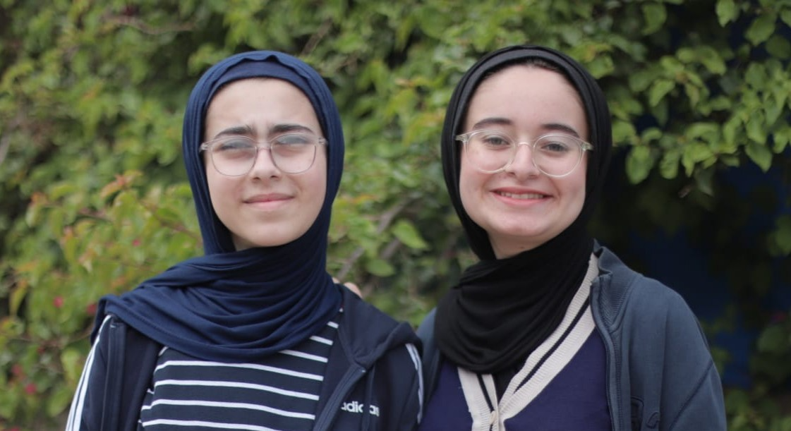 Tala y Farah Mousa, dos chicas adolescentes. Ambas llevan la cabeza cubierta con hiyab, gafas y chaquetas azul marino con cremallera, y sonríen a la cámara frente a un gran árbol verde.