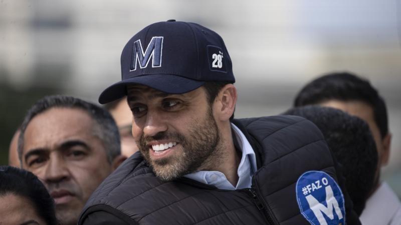 Pablo Marçal sorrindo e olhando para o lado, com boné e adesivo de campanha, rodeado por outras pessoas