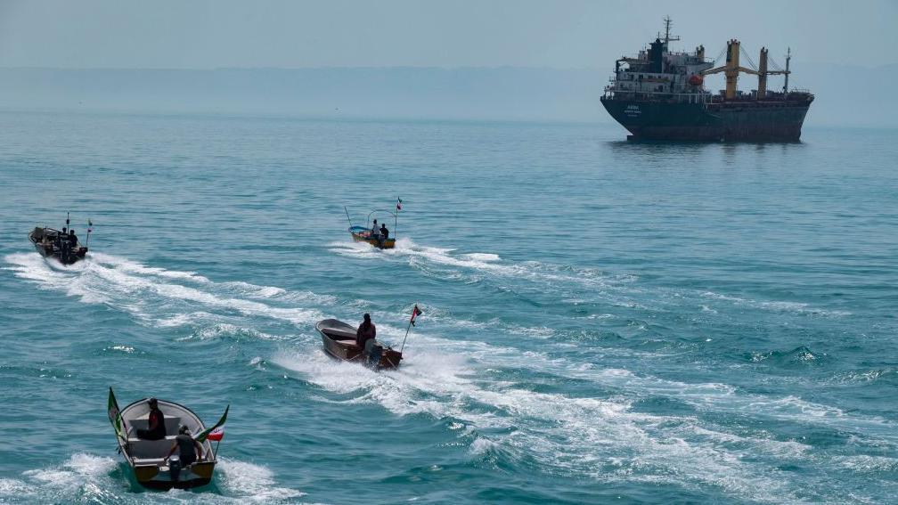 Cuatro lanchas rápidas con banderas iraníes navegando por el Golfo Pérsico, cerca de un buque de carga general.
