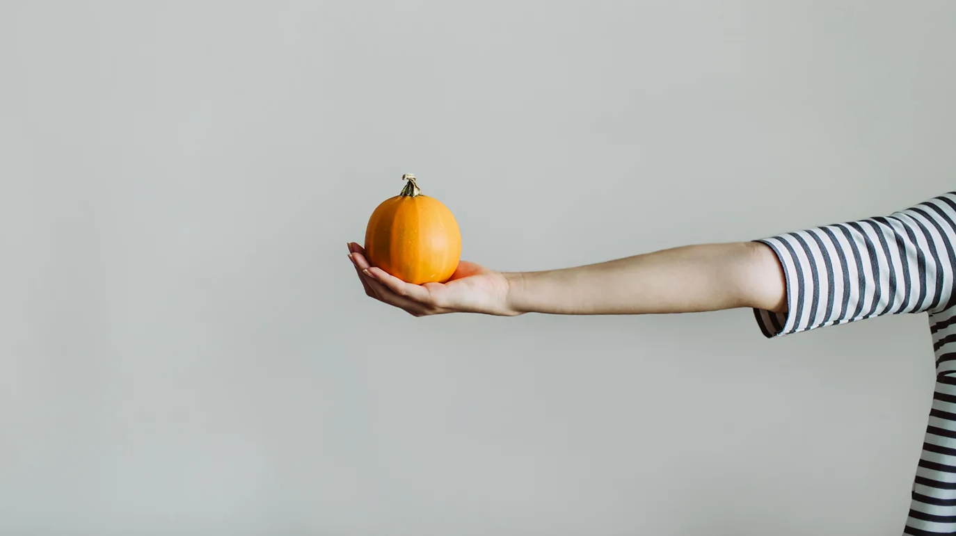 회색 배경 앞에서 팔을 뻗은 사람이 손바닥 위에 작은 주황색 호박을 올려 놓고 있다