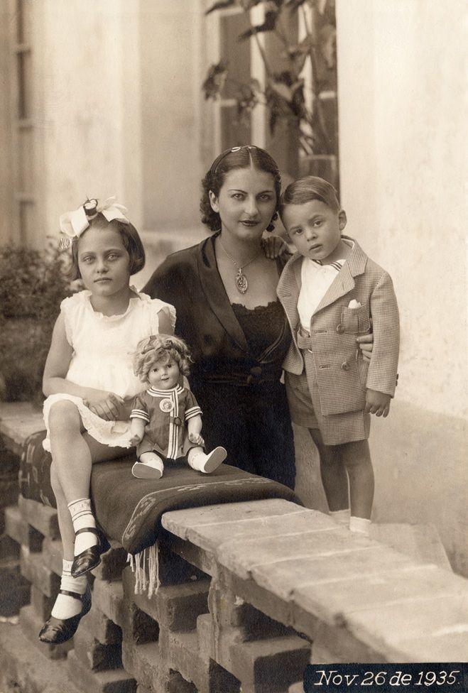 Cristina, en el centro, lleva un vestido y una blusa negros y un collar. A su lado está Antonio, quien lleva una camisa blanca y una chaqueta y un pantalón corto color gris. Al otro lado, sentada, está Isolda, que viste un traje blanco sin mangas, medias blancas y zapatos negros y un lazo en la cabeza. Una muñeca está sentada delante de ellas. Están en un jardín.
