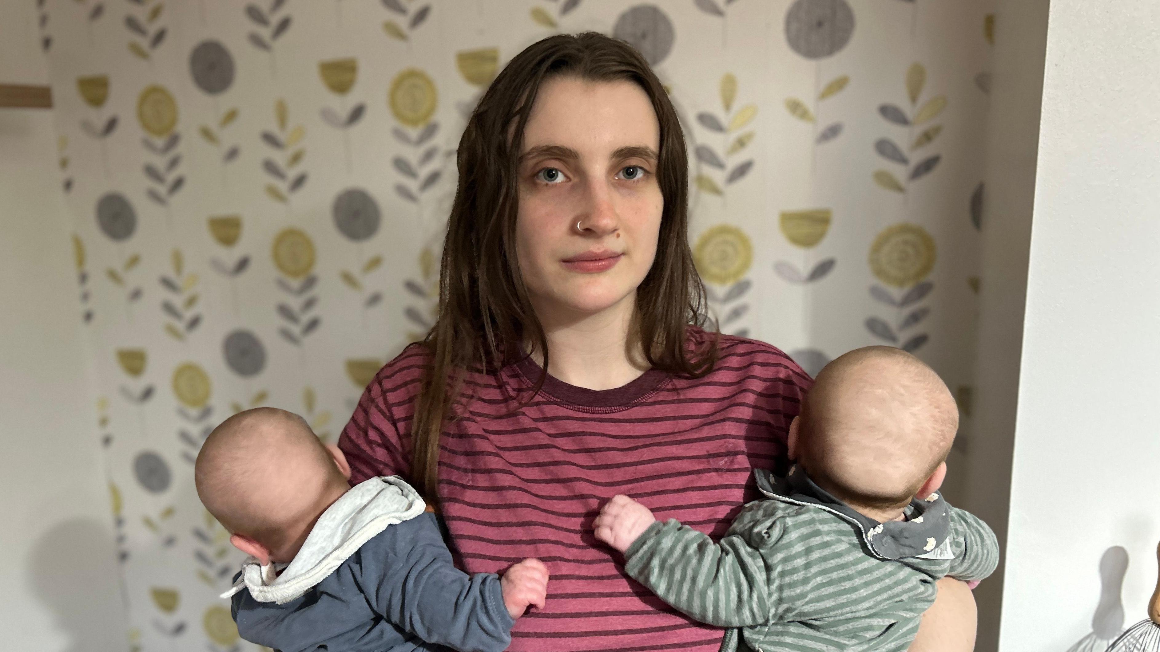 Ella Marcham tem cabelos castanho-escuros e olha para a câmera enquanto segura dois bebês; ela usa uma camiseta listrada vermelha, e os bebês vestem azul-marinho e verde
