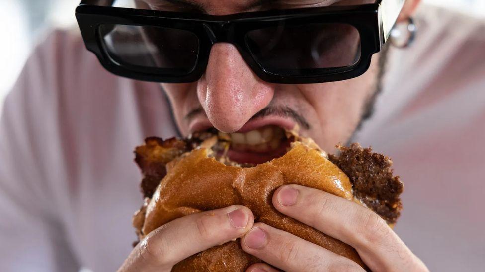 Čovek sa naočarima za sunce grize hamburger koji drži u rukama