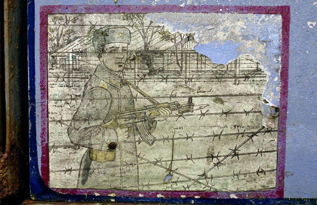 Mural desconchado instructivo para la vigilancia de campos de prisioneros que  muestra a un soldado de pie junto al alambre de púas de un gulag genérico, sosteniendo su fusil AK-47 y vestido con gorro de piel y uniforme de la época.