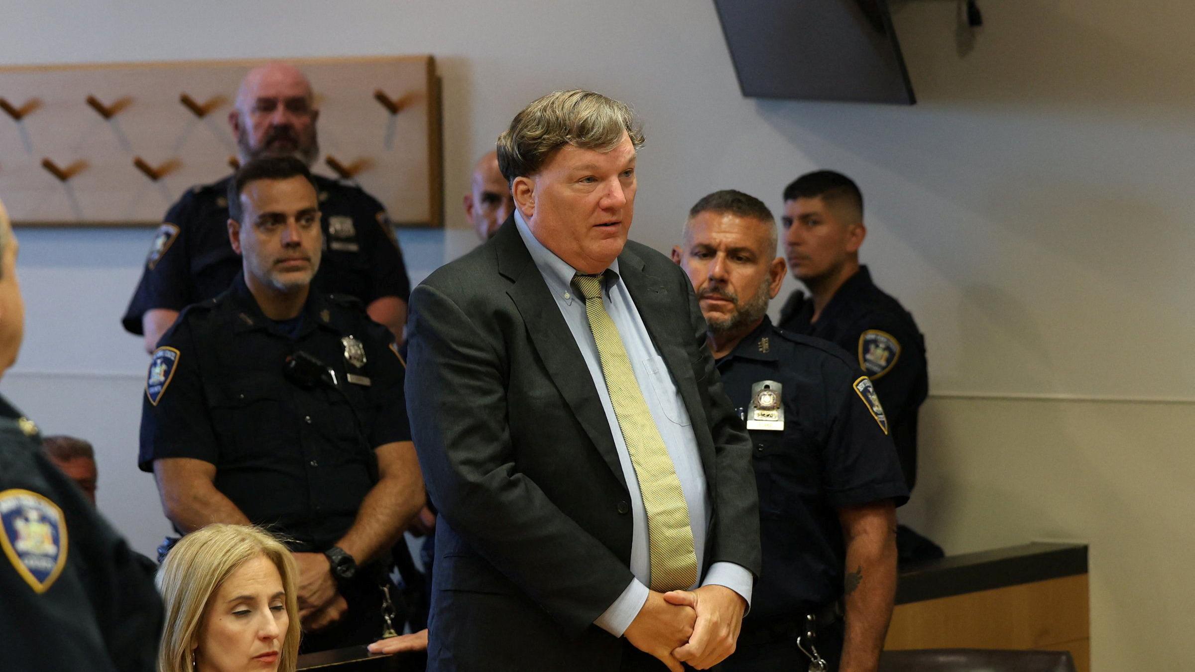 Rex Heuermann, con traje oscuro y camisa y corbata claras en el tribunal, con las manos entrelazadas y gesto serio, rodeado de agentes de policía y una mujer sentada a su derecha