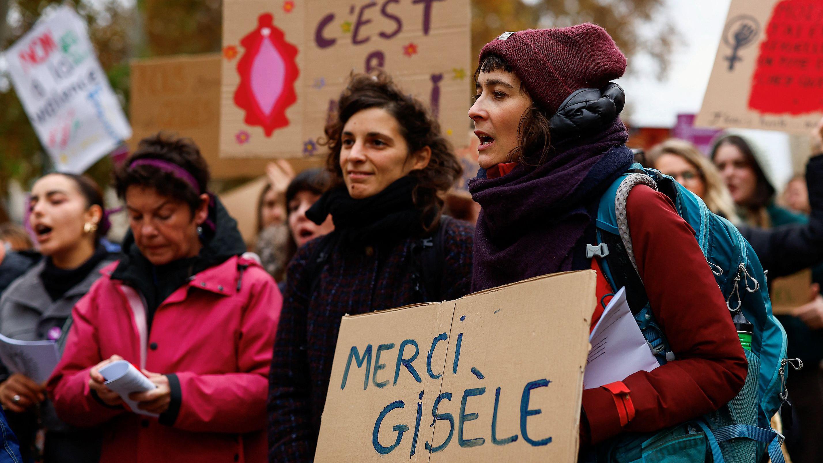 Mulheres em frente ao tribunal de Avignon, segurando cartazes em apoio a Gisèle Pelicot. Uma delas, na frente da imagem, segura uma placa de papelão escrita à mão com os dizeres em francês: 