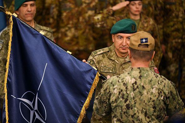 Ulutaş'ın yüzü görünüyor, önde arkası dönük kamuflaj içinde bir asker daha var. En solda nato bayrağı görünüyor.