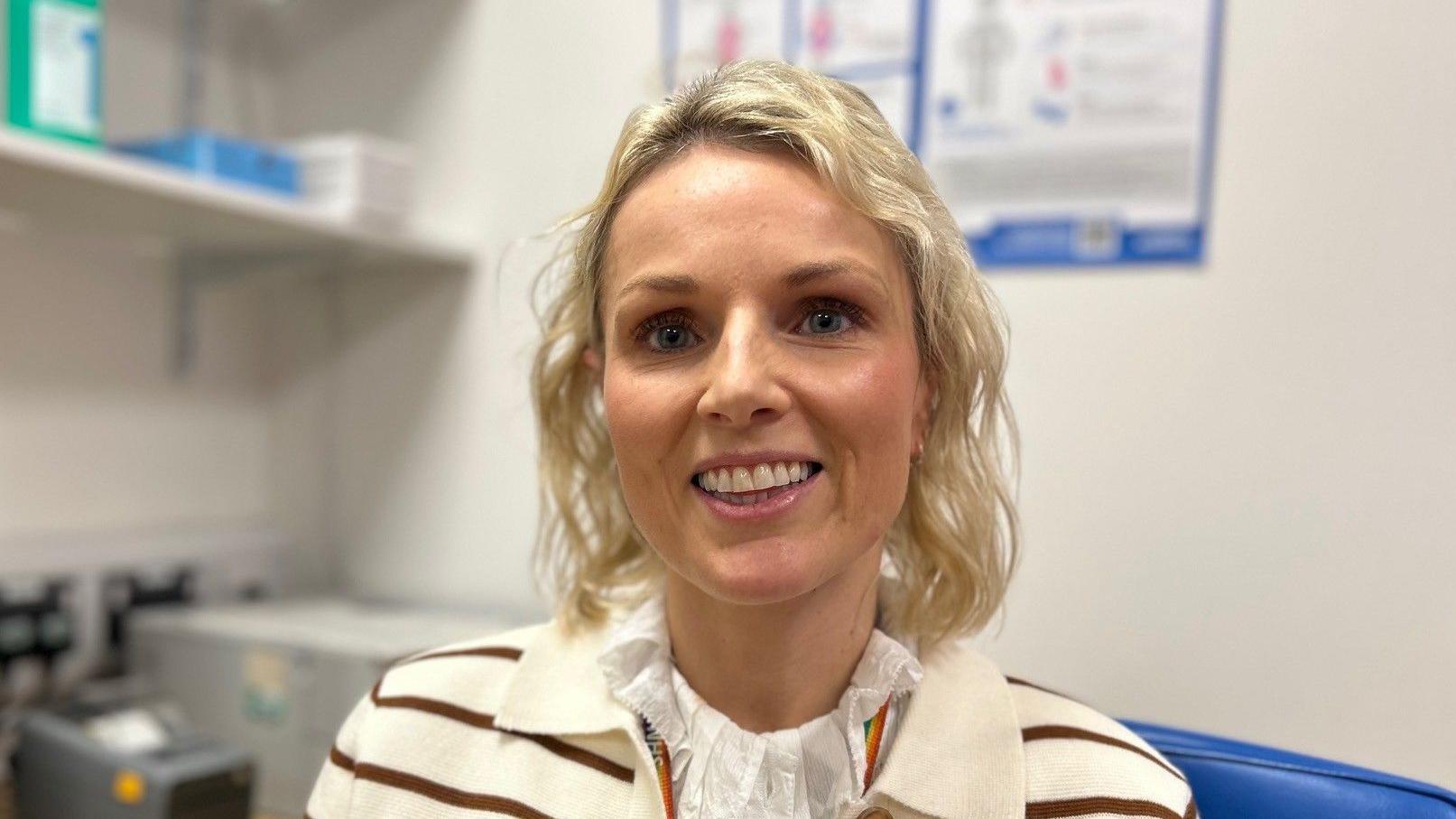 Dr Gilmore has blonde, shoulder length curly hair. She is wearing a brown and white striped cardigan and white blouse. She is sitting in her consulting room.