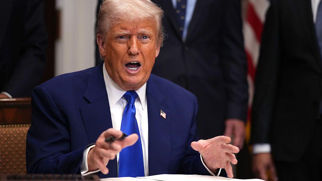 Donald Trump hablando con vehemencia en una firma de una orden ejecutiva. Viste un traje azul oscuro, camisa blanca y corbata azul. Y tiene un pin con la bandera de Estados Unidos.