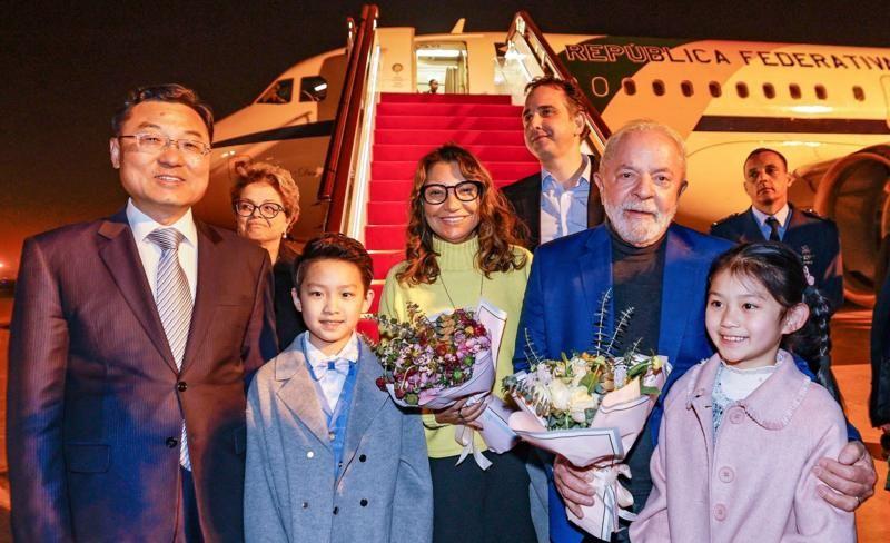 À noite, casal presidencial sorri para foto ao lado de crianças e autoridades chinesas que os receberam com flores na saída do avião, que é visto ao fundo da foto