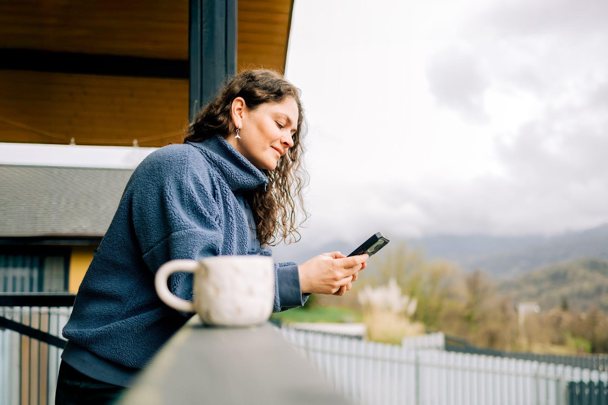 Mulher apreciando o caf&eacute; da manh&atilde; e usando o celular na varanda, com vista para a natureza.