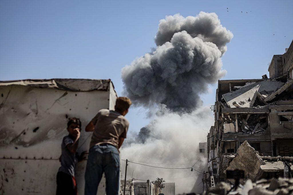 طفلان من غزة يقفان خلف ما يبدو أنه خيمة، يشاهدان أعمدة الدخان المتصاعدة من القصف وأمامهما مبنى مدمر، في صورة التقطت نهاراً.