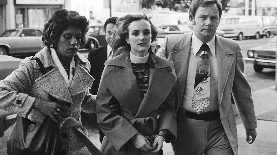 Patty Hearst, una joven rubia vestida con un buzo cuello tortuga, un sweater y una gabardina, siendo escoltada por dos agentes del FBI, una mujer y un hombre.