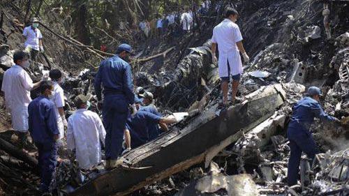 En medio de vegetación, el fuselaje de un avión destruido tras lo que parece ser un accidente aéreo. Alrededor, una decena de hombres vestidos con uniformes observan la escena.