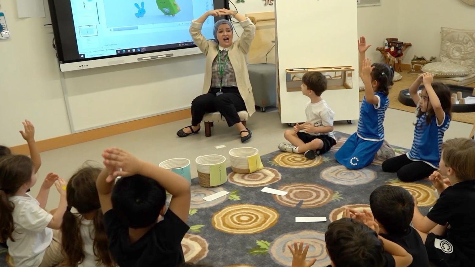 صورة تُظهر صفّاً فيه عددٌ من الطلاب الصغار وهم يرفعون أيديهم فوق رؤوسهم والمعلمة تجلس قبالتهم، في نشاط غنائي لتعلّم اللغة العربية.