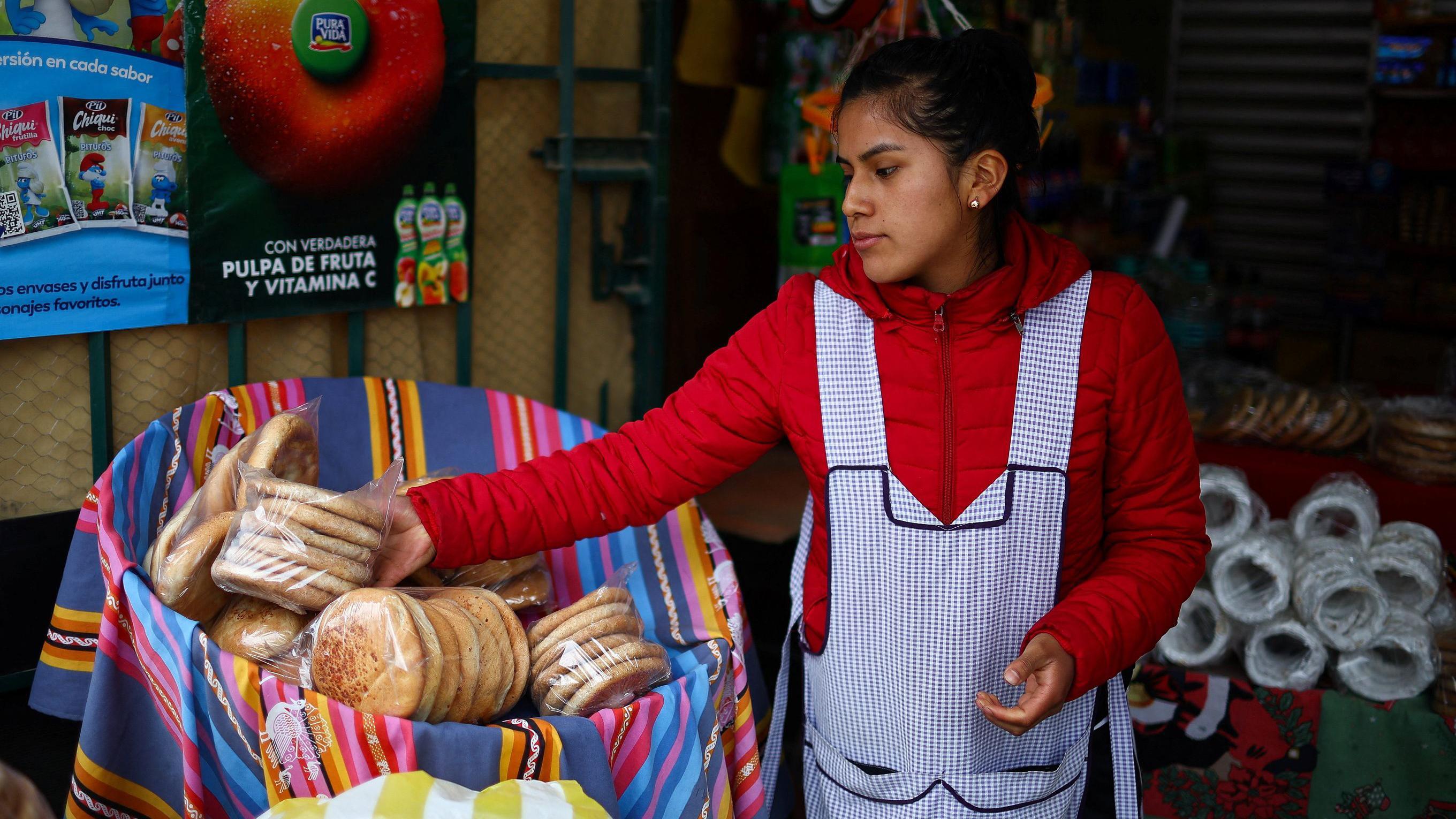 Una mujer en una tienda, coloca lo que parece ser pan dulce en un canasto. Viste una chaqueta roja y tiene un delantal a cuadritos en tono azul y blanco