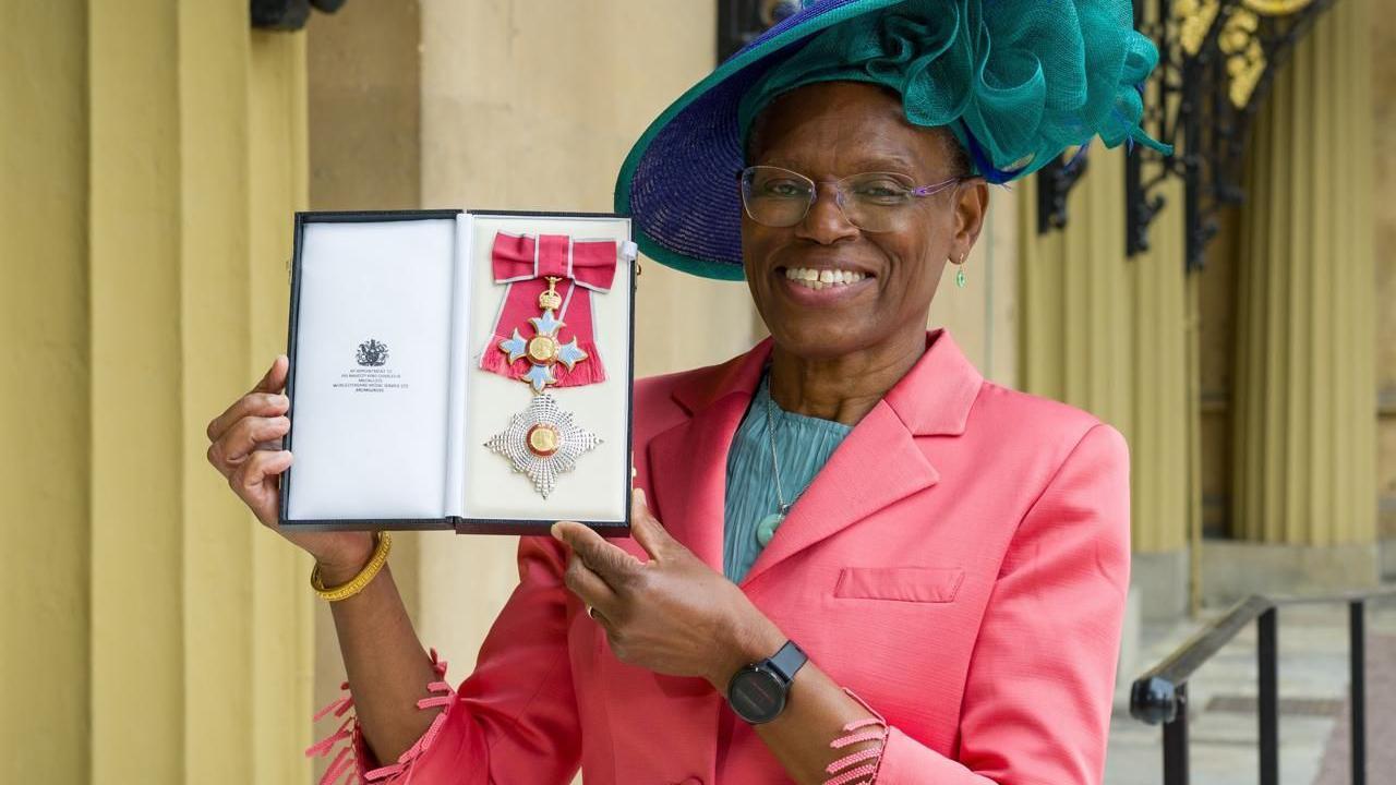 Ijeoma Uchegbu posa en primer plano vestida con una chaqueta de color coral y una blusa celeste, además de con un sombrero de ala de lado, también celeste. Sonríe. Lleva en la mano un estuche largo, abierto, donde está la condecoración de Dama Comandante de la Más Excelente Orden del Imperio Británico. La condecoración es una gran lazo de color coral con ribetes plateados y una larga cruz dorada, plateada y celeste.