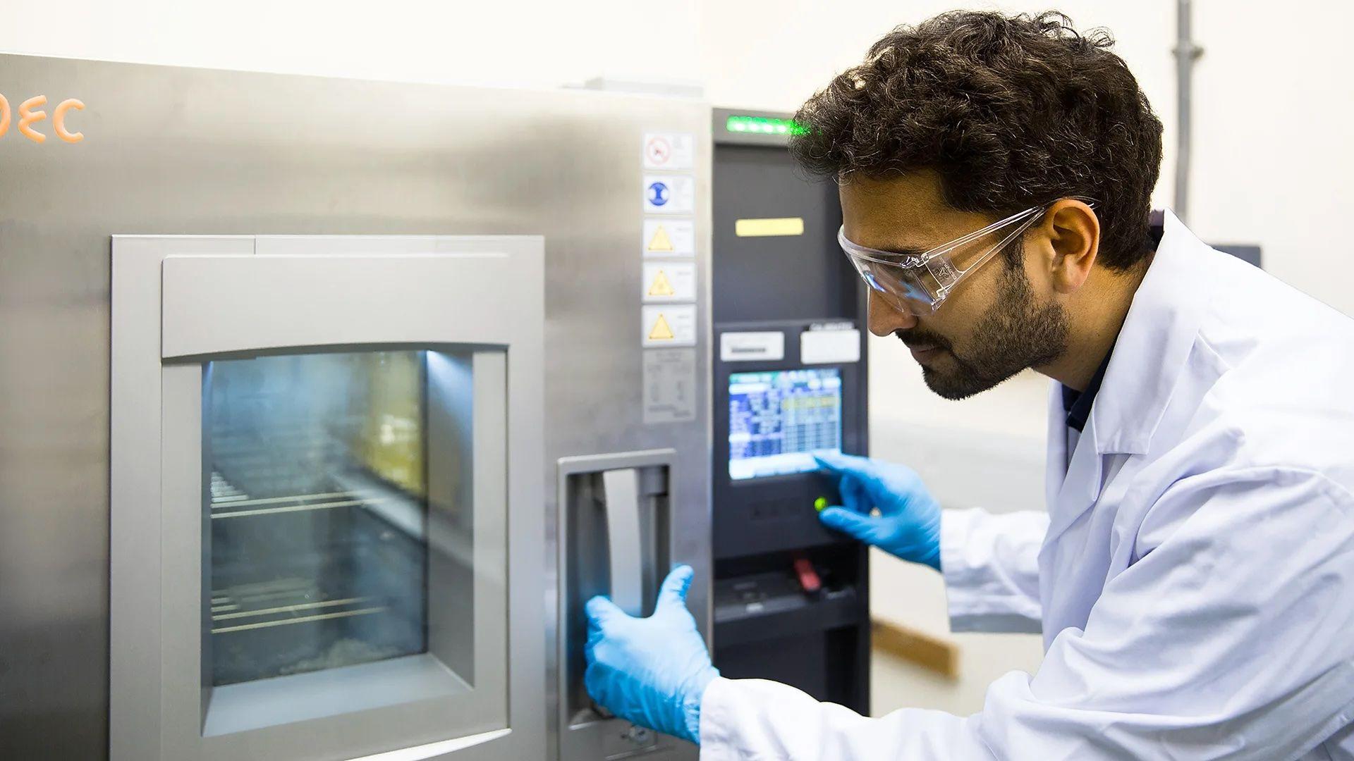 Un investigador con bata blanca, guantes de látex y gafas protectoras opera una especie de horno en el que se encuentran paneles solares.