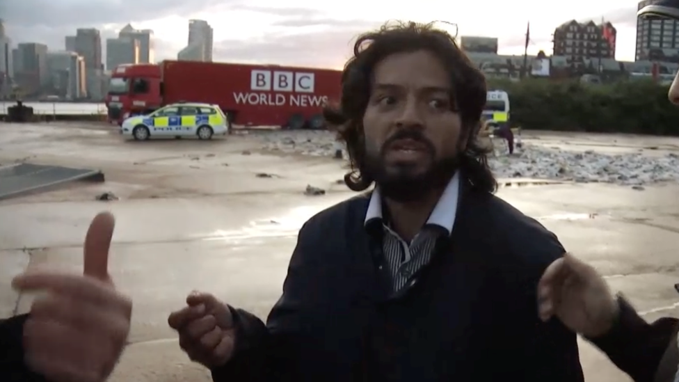 Um homem com longos cabelos escuros, barba e vestindo uma jaqueta está com um carro de polícia falso e um caminhão falso da BBC World News ao fundo.