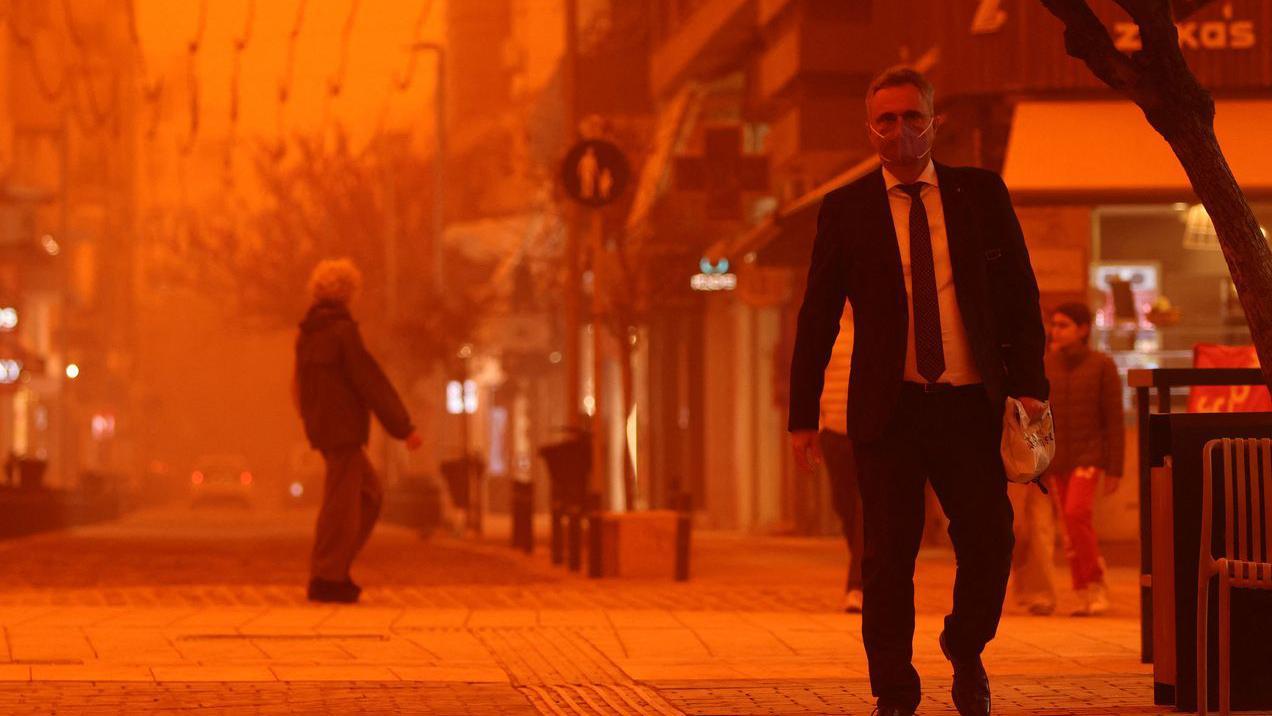 Čovek u odelu sa maskom za lice drži papirnu kesu u ruci, sve oko njega je narandžasto, u daljini se nazire stariji muškarac