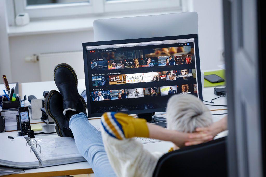 Una persona mira un catálogo de series en un monitor, mientras se sienta relajado con los pies en el escritorio.