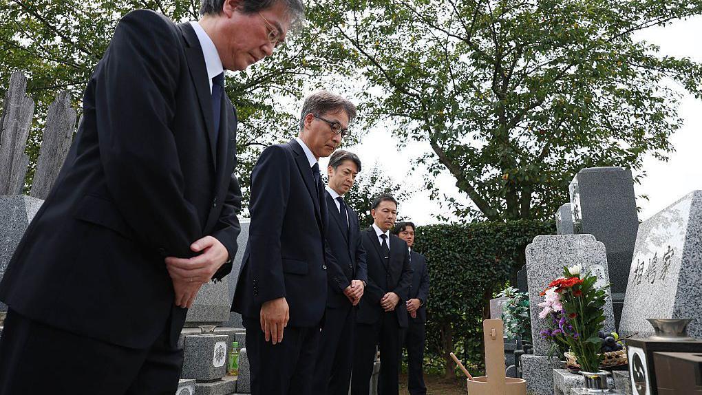 Pet muškaraca u tamnim odelima klanja se pred grobom u Japanu. Grob je napravljen od sivog mermera i ispred njega se nalazi vaza sa cvećem.