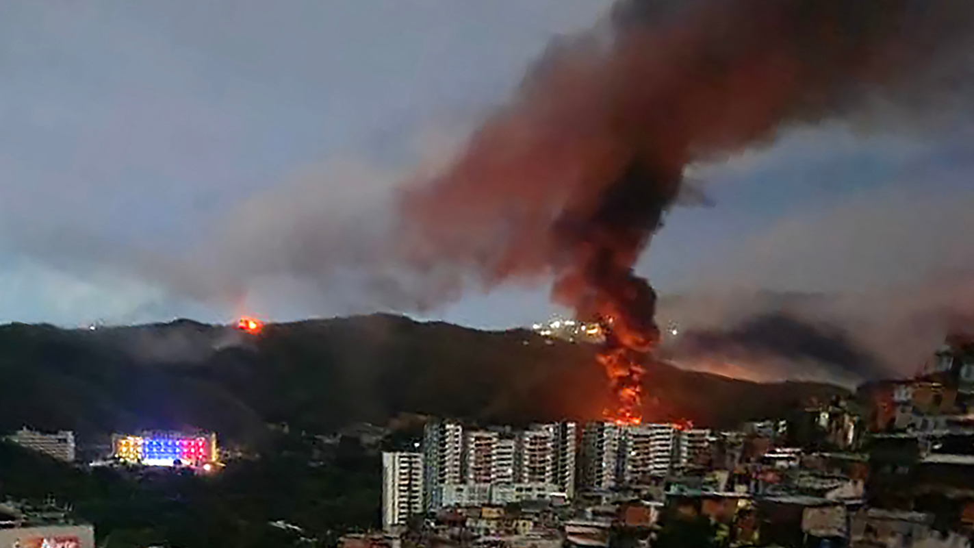 Una nube de humo a lo lejos en medio de la ciudad de Caracas, donde se ven viviendas y edificios y una montaña