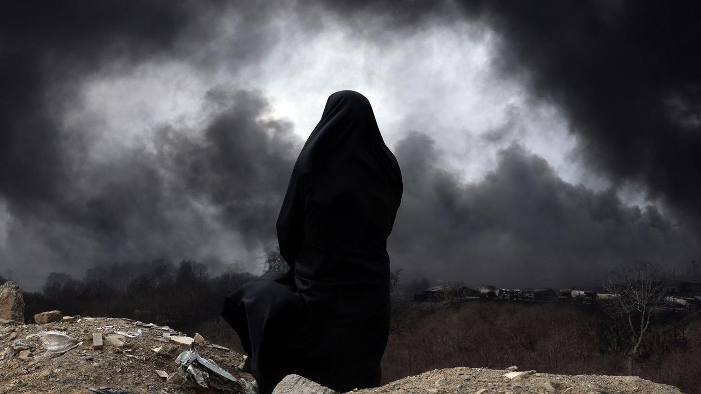 Vista pelas costas, uma mulher usando um chador observa a paisagem enquanto uma fuma&ccedil;a preta se espalha pelo horizonte