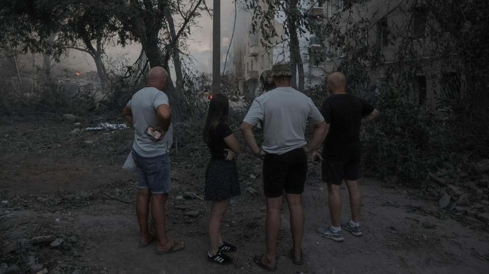 Três homens de shorts e camiseta e uma jovem com roupas leves, de pé em um jardim, observando a fumaça que sai dos destroços causados por um ataque em Kramatorsk, atribuído ao exército russo.