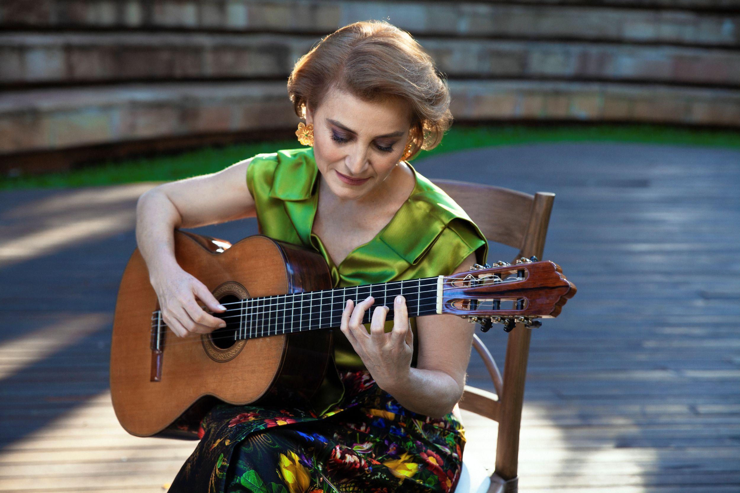 Berta Rojas toca su guitarra. Está sentada en una silla de madera.