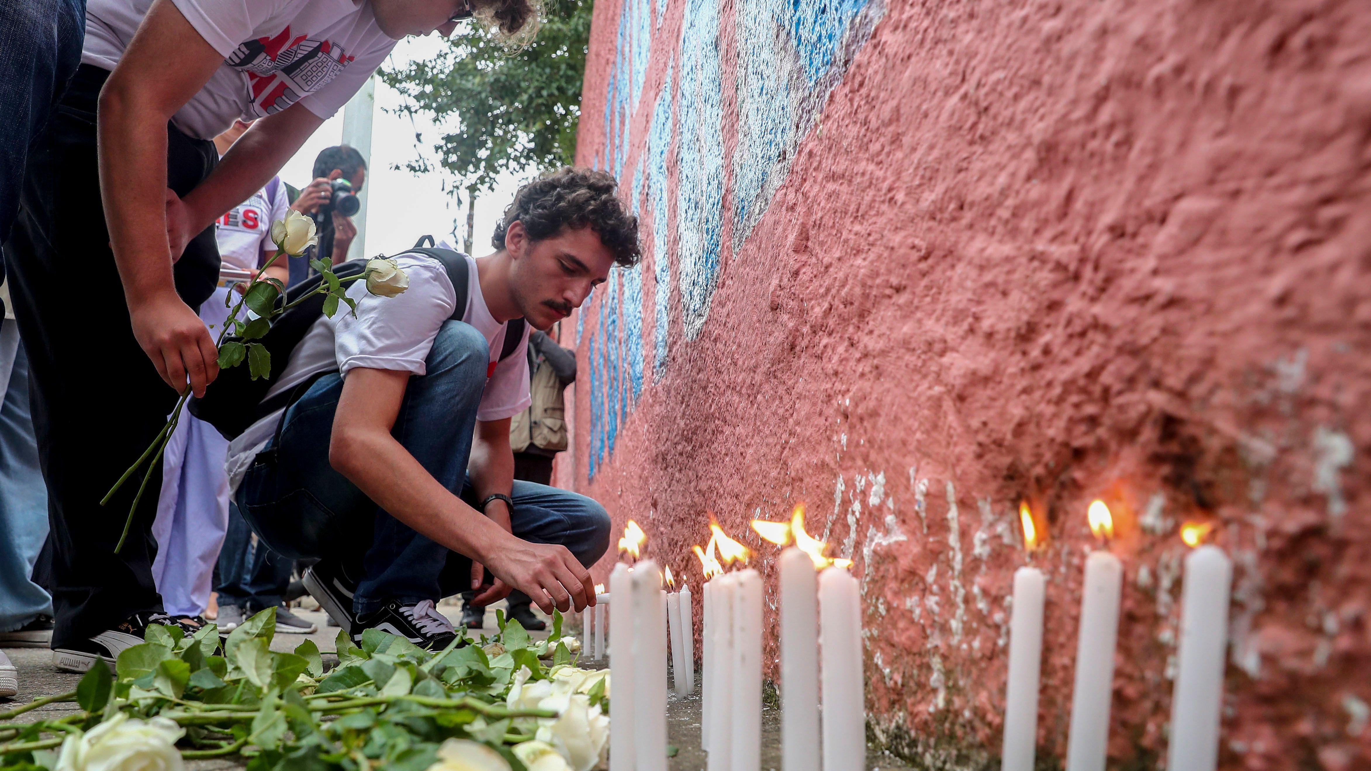 Velas e flores são deixadas em muro em frente a escola em São Paulo, no fundo um homem de camisa branca acende uma vela e a mão de uma mulher segura uma flor