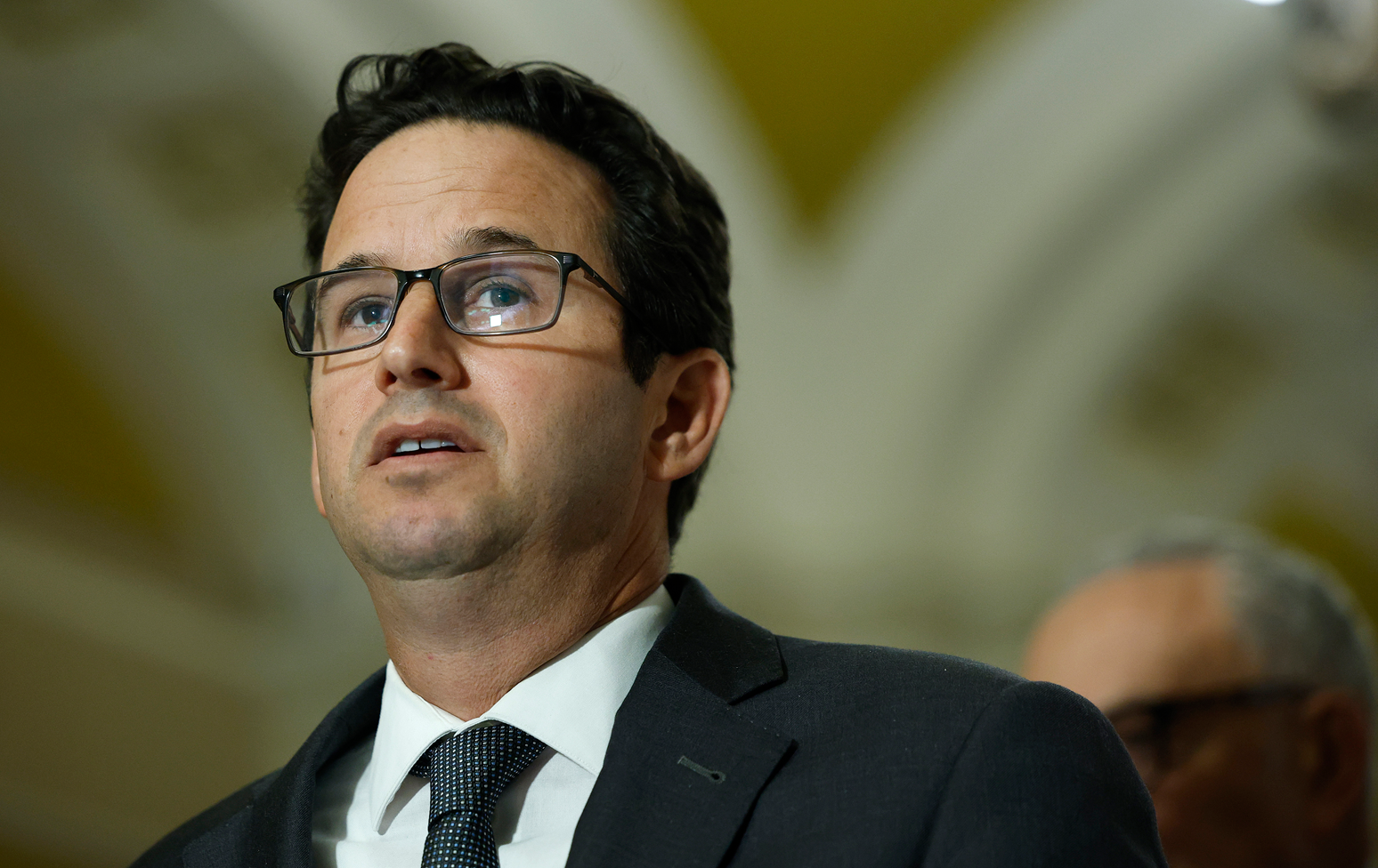 Brian Schatz con lentes, una camisa blanca, chaqueta negra y una corbata negra con puntos blancos