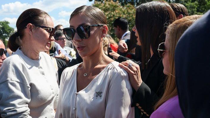 Marina Lacerda em p&eacute;, rodeada por outras mulheres; ela tem olhar s&eacute;rio, de quem esta observando algo