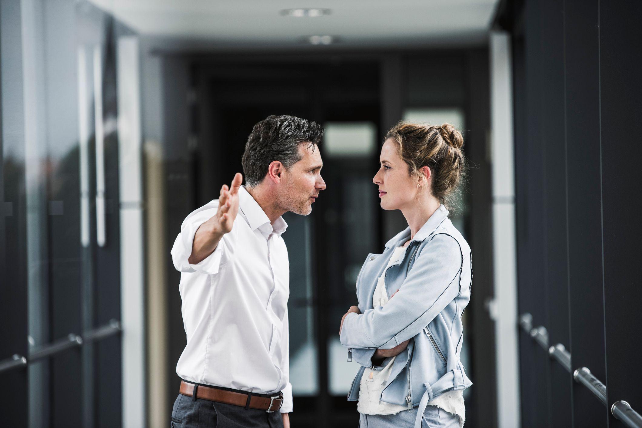 Um homem e uma mulher, num corredor de uma empresa, discutem. Ela encara seriamente para ele, que est&aacute; apontando para frente 