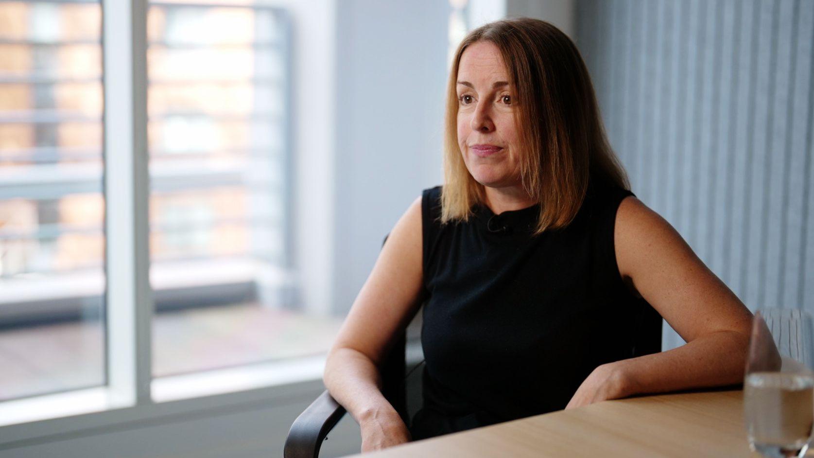 Tessa Gregory sentada en una silla, junto a una ventana y vestida de negro