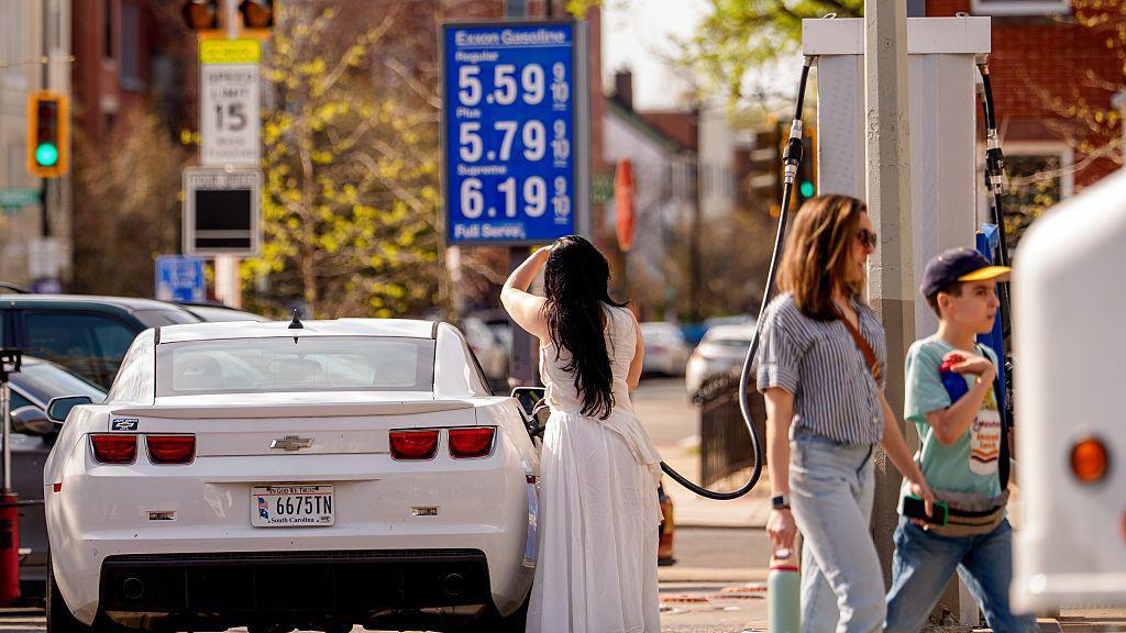 Uma mulher de vestido branco est&aacute; no centro da imagem. Ela abastece um carro branco em um posto de gasolina em Washington D.C., EUA

