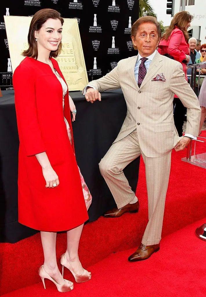 Anne Hathaway vestindo um casaco vermelho at&eacute; a canela e salto alto posa com Valentino em um tapete vermelho. 