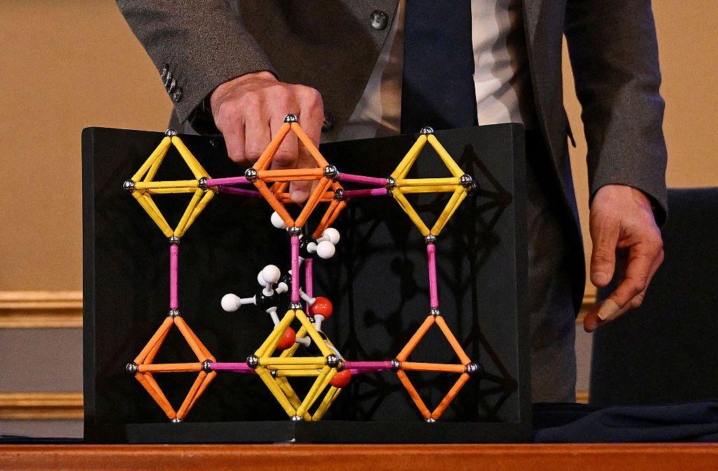 En primer plano se ve un modelo de colores que trata de mostrar la estructura molecular que contiene uno de los MOF. Tienen detrás una plataforma oscura para poder destacar los colores del modelo. 