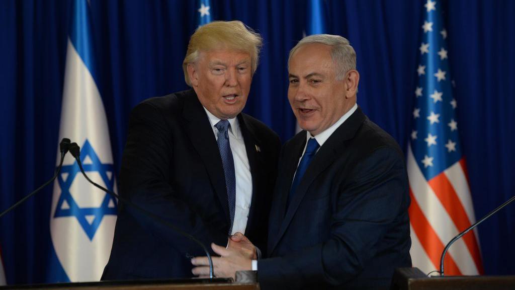 Američki predsednik Donald Tramp i izraelski premijer Benjamin Netanjahu rukuju se ispred izraelske i američke zastave u Jerusalimu 22. maja 2017. Obojica su obučeni u tamna odela sa plavim kravatama.